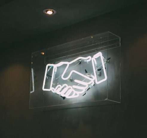 On a black wall there is a white neon sign of two hands shaking. Aligned teams can reach peak performance. Image for peak performance blog.